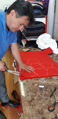 A man holding scissors to a piece of red material with mask shapes drawn on it.